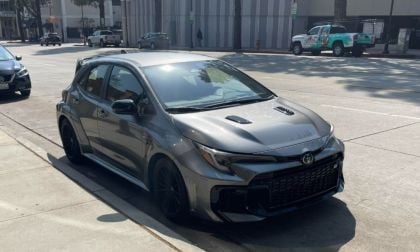 A dark gray Toyota GR Corolla is parked on a sunlit street. Its shiny exterior and sporty design convey a modern, dynamic feel. Nearby cars and city buildings form the backdrop.