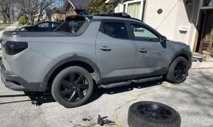 A grey Hyundai Santa Cruz with black wheels is parked in a driveway. One wheel is removed lying beside the truck. Sunlight casts shadows; tools are on the ground.