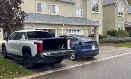 Blue Tesla Model Y being charged from white GMC pickup truck's bed outlet, rear three-quarter view, residential driveway setting.