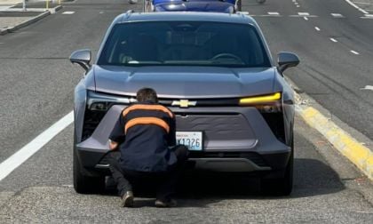 A person crouches in front of a silver Chevrolet Blazer EV on a road. They are focused on the front grille, with a road and another vehicle visible behind.