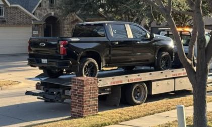 A black Chevrolet Silverado pickup truck being loaded onto a flatbed tow truck, photographed from the side angle in a residential driveway.