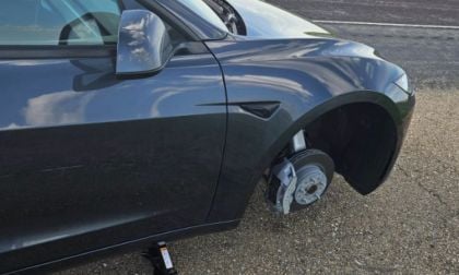 A black Tesla Model 3 parked roadside with front wheel missing, revealing the brake rotor and caliper. A car jack is also positioned beneath the vehicle.