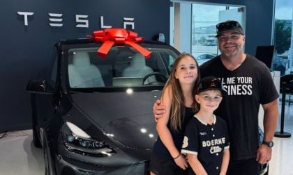 Clint's family stands in front of a black Tesla Model Y with a large red bow on top. They smile warmly in a Tesla showroom, conveying joy and excitement.