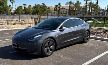 Sleek gray Tesla Model 3 parked in desert residential area, angled side view with palm trees and metal fence background