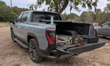 A gray GMC Sierra EV Denali parked on dirt with its tailgate down, carrying a chainsaw, toolbox, gloves, a drink can, and a water bottle on the bed