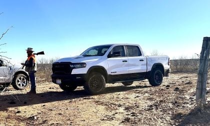 White 2026 Ram 1500 Rebel parked on a dirt road under a clear blue sky. Nearby, a man in an orange vest and hat stands beside another vehicle. Sparse desert surroundings.