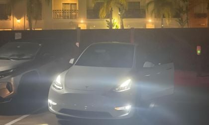 White Tesla Model Y with headlights on, parked in dimly lit lot with palm trees and building lights in background