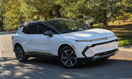 A white Chevrolet Equinox EV shown from a front three-quarter view, featuring black accents and distinctive two-tone wheels, parked in a shaded outdoor setting.
