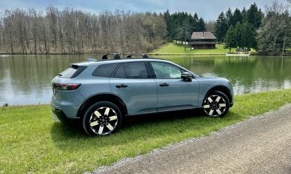 A sleek, light blue 2024 Honda Prologue with shiny black and white rims is parked by a tranquil lakeside. Bare trees and a rustic cabin create a peaceful, scenic backdrop.