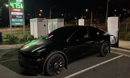 Black Tesla Model Y plugged into a Tesla Supercharger at night with nearby gas prices displayed in the background.