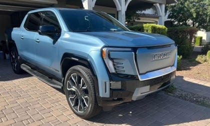 Light blue GMC Sierra Denali Ultimate pickup truck parked on brick driveway with landscaped background, showing bold front grille and premium styling