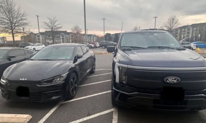 A black 2023 Kia EV6 and a dark blue Ford F-150 Lightning are parked side-by-side in a parking lot, both shown from their front three-quarter angles at dusk with apartment buildings visible in the background.