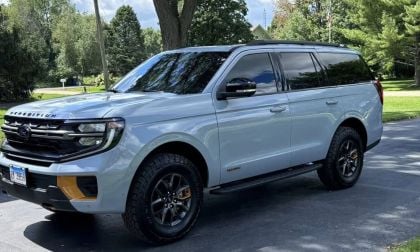 Elegant Grey Ford Expedition Tremor parked in a suburban driveway. The sun casts reflections on its sleek exterior. Green trees and a well-kept lawn surround the scene.