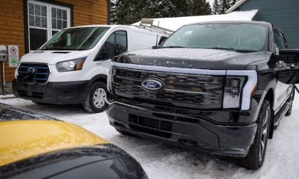 A black Ford F-150 truck and a white Ford Transit van parked in a snowy area near a wooden building, with charging stations visible.