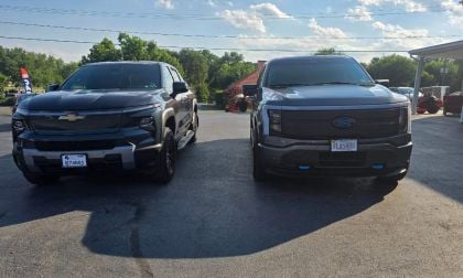 Black Chevy Silverado and Ford F-150 Lightning electric trucks parked side-by-side on asphalt lot with trees and blue sky background