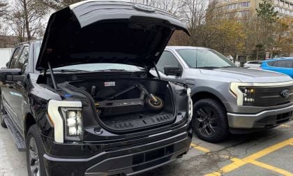 A black electric truck with an open front trunk displaying a scooter sits beside a gray electric truck in a parking lot.