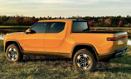Orange Rivian R1S electric SUV shown from the rear three-quarter view, parked on grass near a lake with autumn trees in the background during sunset.