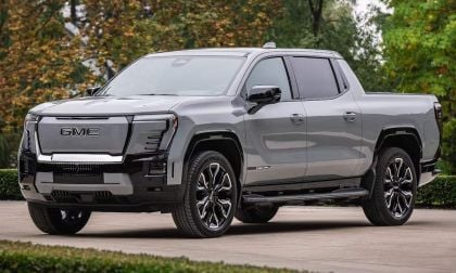 A gray 2025 GMC Sierra EV pickup truck parked outdoors, showcasing its modern design and bold front grille against a blurred natural backdrop.