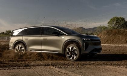 A silver 2025 Lucid Gravity drives through dirt, kicking up dust, with mountains in the background under a clear blue sky.