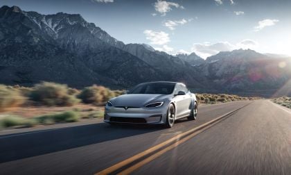 A gray 2025 Tesla Model S drives along a winding road with majestic mountains in the background under a bright sky.