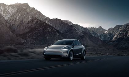 A sleek electric car drives on a deserted road with dramatic mountain peaks in the background, illuminated by soft sunset light.