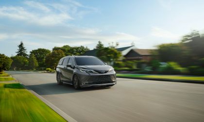 A dark gray 2024 Toyota Sienna minivan shown from the front three-quarter angle, driving on a suburban road with motion blur effect and green landscaping in the background.