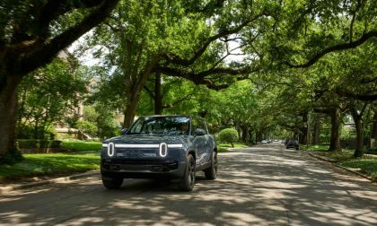 A blue 2025 Rivian R1S SUV drives down a tree-lined street, showcasing modern design amidst lush greenery and suburban homes.