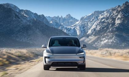 A 2025 Gray Tesla Model Y drives on a rural road, set against a backdrop of snowy mountains under a clear blue sky.