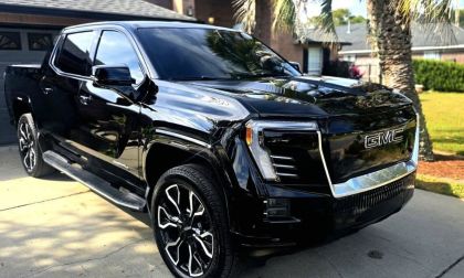 A black GMC Sierra EV shown from a front three-quarter angle parked in a residential driveway, featuring distinctive black and chrome wheels and LED lighting elements.