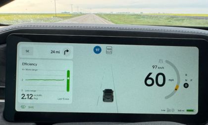 Rivian R1T dashboard showing a digital display. Speed is 60 mph, energy use is 2.12 mi/kWh, with 24 miles to the next turn. Background shows open road with luscious flat-land greenery.