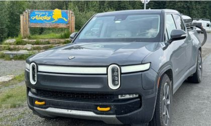 Gray Rivian R1T electric pickup truck parked by Alaska welcome sign, surrounded by forest landscape