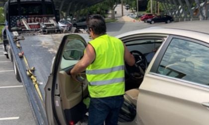A man in a yellow safety vest prepares to drive a beige Hyundai Sonata onto a tow truck. The scene is in a parking lot with a few parked cars nearby.