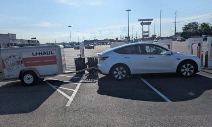 A white Tesla Model Y is parked at a charging station while towing a U-Haul trailer. The scene is in a parking lot under a clear blue sky, conveying a sense of travel.