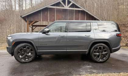 A sleek, gray 2026 Rivian R1S is parked on a wet driveway in front of a rustic wooden structure. Bare trees and a calm, overcast sky create a serene backdrop.