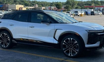 A white Chevrolet Blazer EV is shown from a front three-quarter angle in a parking lot, featuring distinctive black accents and two-tone alloy wheels.