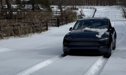 A black Tesla Model Y is parked in a snow-covered driveway, photographed from a three-quarter front angle with winter fencing and trees visible in the background.