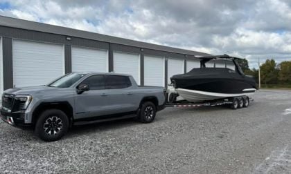 A grey GMC Sierra EV parked on gravel in front of storage units, towing a large black boat with a triple-axle trailer. The scene is under a partly cloudy sky.
