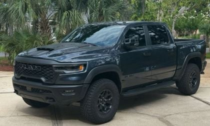 A black Ram 1500 RHO pickup truck is parked on a driveway. The truck features a bold front grille and rugged tires. Lush greenery and trees are in the background.