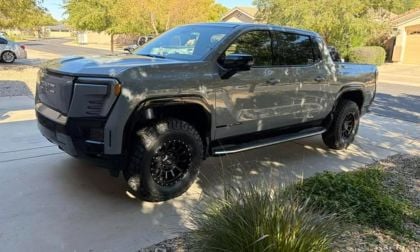 Gray GMC Sierra EV with black off-road wheels, lifted suspension, and custom front bumper parked on residential driveway in suburban setting.