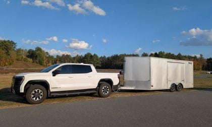 A white GMC Sierra EV is parked on a paved road, towing a large white enclosed trailer. The background features a grassy field and a clear, sunny sky.