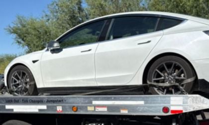 A white Tesla Model 3 is secured on a flatbed tow truck in a sunny outdoor setting with trees in the background, suggesting it needed roadside assistance.