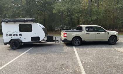 A white Ford Maverick is parked in a forested area, towing a small teardrop camper with "APPLACHIAN" printed on its side. The scene is calm and outdoorsy.