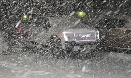 A GMC Sierra EV photographed during heavy snowfall at night, with its illuminated GMC badge visible through the swirling snow and yellow lights glowing in the background.