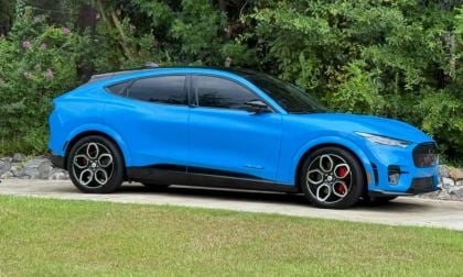 A gleaming, bright blue Ford Mustang Mach-E parked on a driveway, surrounded by lush green foliage. The car's modern design conveys a sense of innovation.