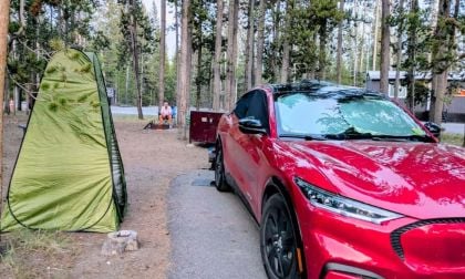 A red Ford Mustang Mach-E is parked in a forest campsite near a green tent. A person sits by a fire pit in the background, surrounded by trees and cabins. The scene feels peaceful and rustic.
