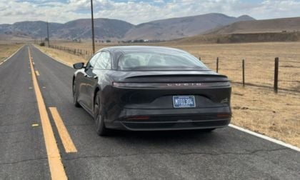 A black Lucid Air drives down an empty rural road flanked by dry fields and distant hills under a partly cloudy sky, conveying a sense of freedom.