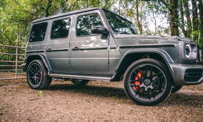A gray Mercedes-Benz G-Class SUV shown from a side-front quarter view, featuring black aftermarket wheels, red brake calipers, and photographed in a wooded setting.