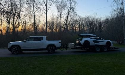 White Rivian R1T towing a trailer with a McLaren at sunset, surrounded by bare winter trees