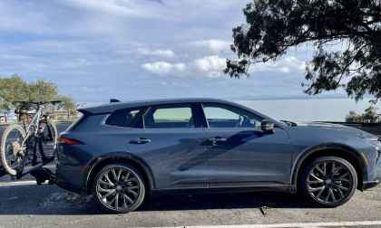 A blue Toyota Crown Signia shown from the side view, featuring black wheels and a bike mounted on a rear rack, parked in an oceanside lot under partly cloudy skies.