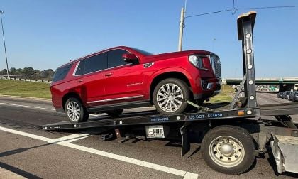 Red GMC Yukon Denali being towed on a flatbed truck on a sunny day. The background shows a highway with light traffic and green grass.